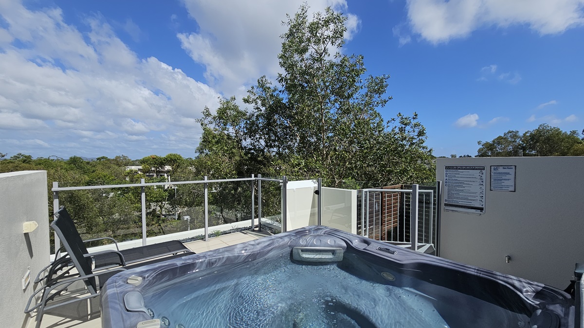 The rooftop spa features a grey hot tub in the middle, flanked by two black sun lounge chairs. Glass railings secure the area. Safety reminder posters are on the right wall. Trees surround the area.