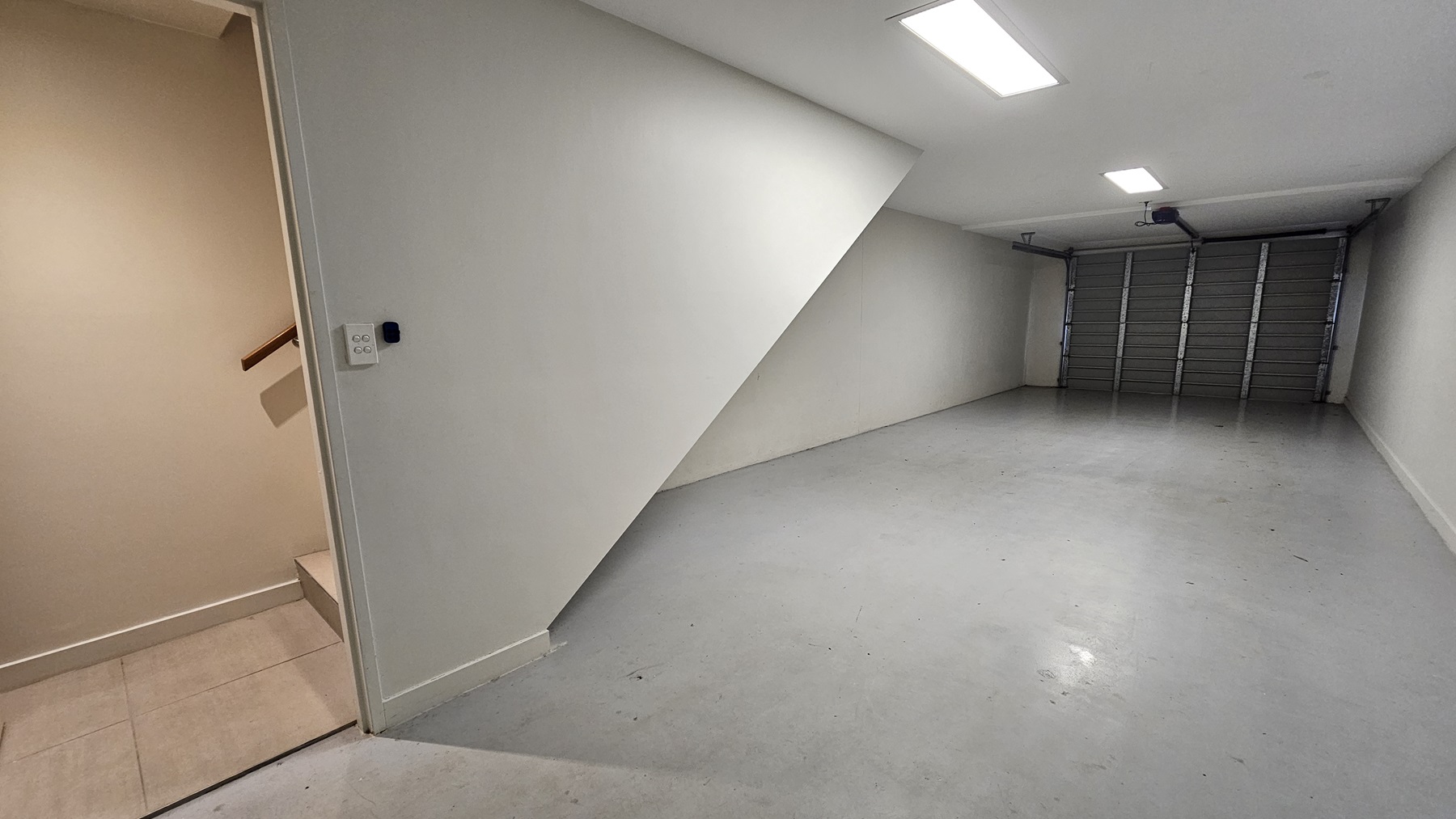 A space with a metal door and hard concrete floor. The white walls and ceiling are illuminated by fluorescent lights. A staircase with handrails on the left grants access to the upper level.