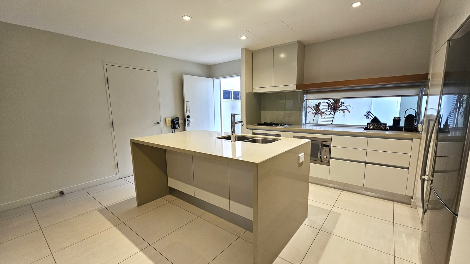 Inside the kitchen, an island bench with cream tiles is seen. Next to it, a large fridge, kitchen cupboards, and equipment are arranged. A phone is mounted on the wall to the left of the door. Cream tiles adorn the floor and ceiling, illuminated by LED lights.