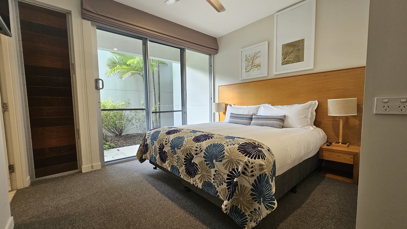 Inside the bedroom, a king bed with printed linens and a wooden headboard is flanked by matching bedside tables and lampshades. Large photo frames adorn the right wall. A glass door on the left leads outdoors, offering a view of small trees. A retractable curtain is affixed to the wall near a wooden door. The floor is covered in grey carpet.