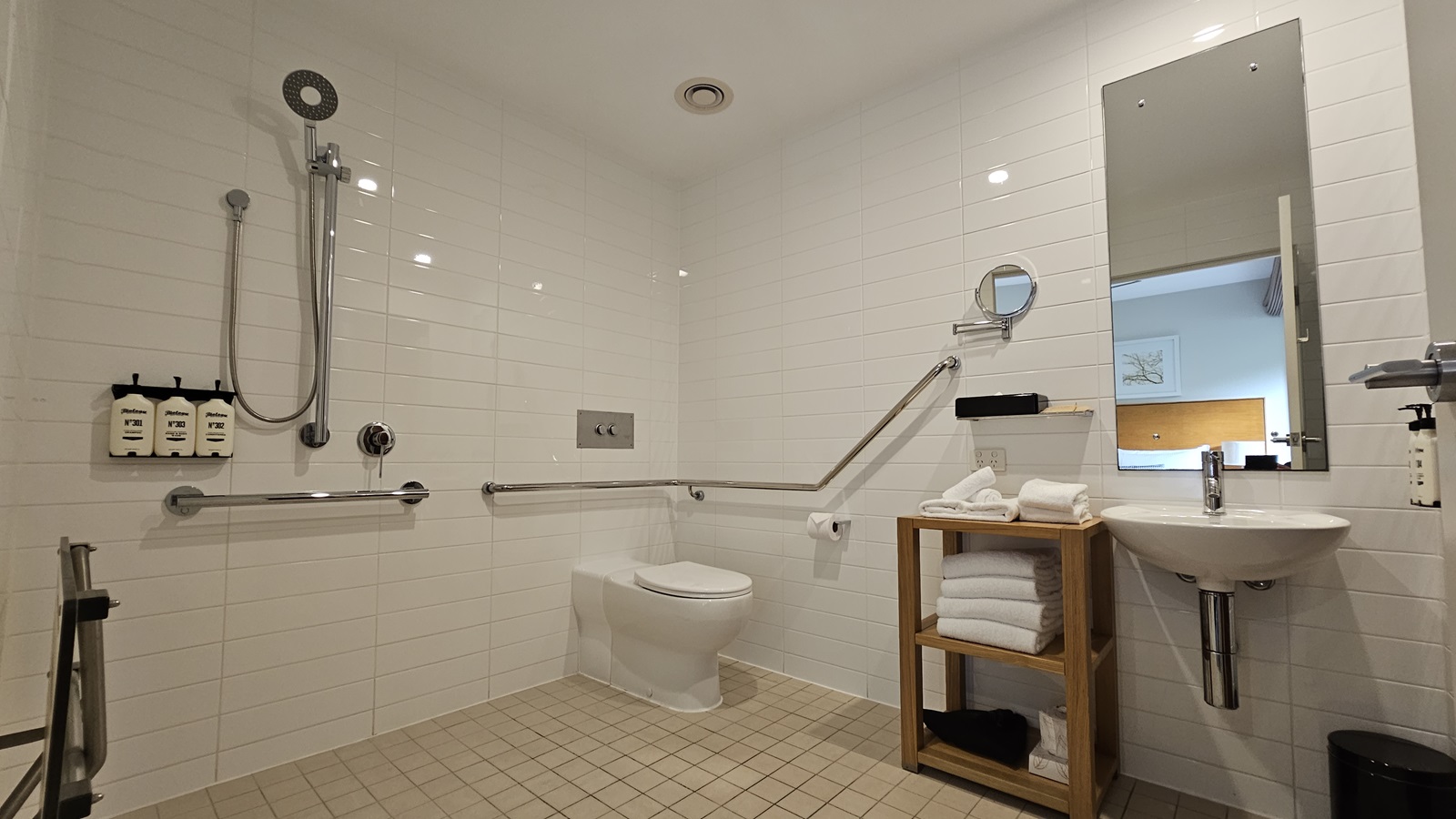 Inside the toilet, a toilet bowl with hand rails in the corner. Tissue dispensers are on the wall, with a shower and handrails to the left. Toiletries are affixed nearby. A wooden storage rack holds folded white towels on the right, beside a mirror, hand basin, and liquid soap dispenser. A rubbish bin is underneath. A foldable bench chair is near the shower. The floor has small cream tiles, while the walls and ceiling are white.