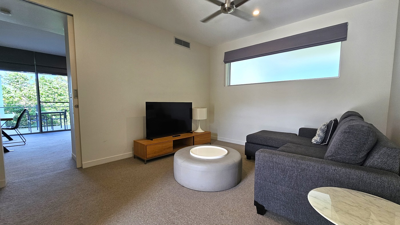 In the corner, a flatscreen TV sits atop a TV stand. A lampshade stands beside it, with a window to its right covered by a retractable curtain. Adjacent is a lounge area with cushioned grey chairs and a round table. The floor has grey carpet, and the wall and ceiling are white, illuminated by an LED light and ceiling fan. To the left is a balcony furnished with tables and chairs.