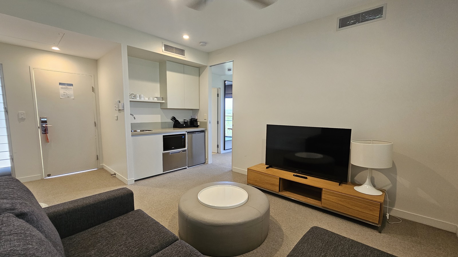 To the right, a flatscreen TV rests on a TV stand, accompanied by a lampshade. Adjacent is a lounge area with cushioned grey chairs and a round table. To the left is a cupboard area with a sink, followed by a door leading outside. The floor has grey carpet, while the wall and ceiling are white, lit by an LED light and ceiling fan.