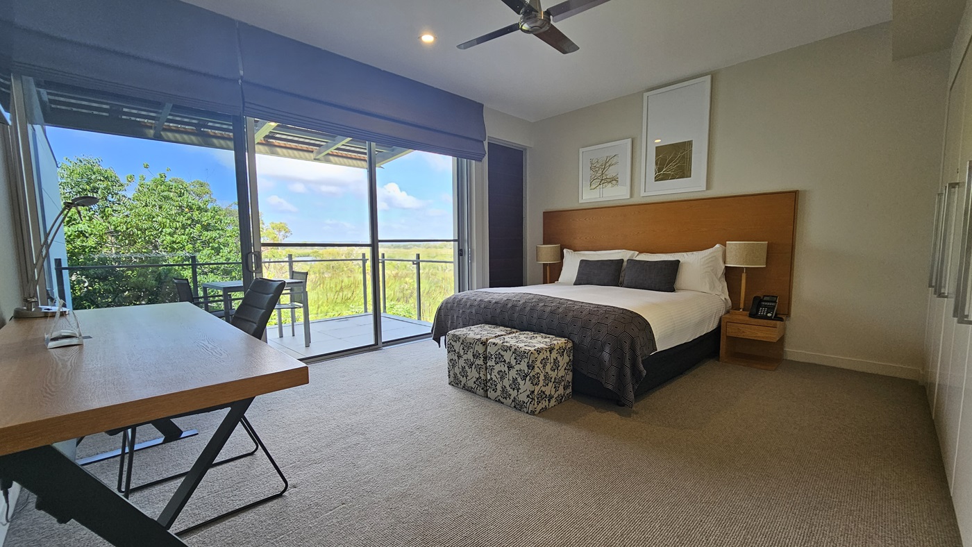 On the right, a king-size bed with a wooden headboard and matching bedside tables. Lampshades top the tables. A glass door to the balcony is on the left. Retractable curtains on the left wall. A wooden office table with a lamp is near the bed. Grey carpeted floor, white walls and ceiling with LED light and ceiling fan. Balcony has tables and chairs.