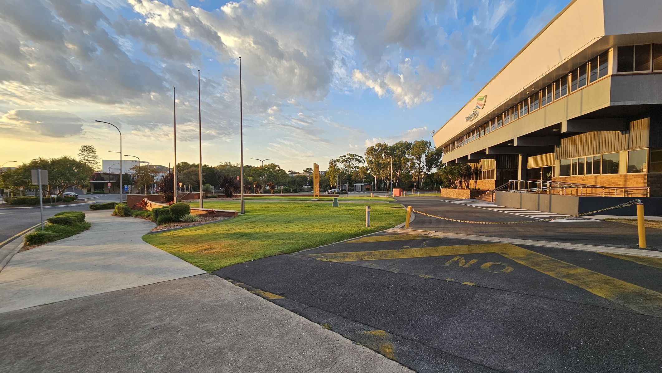 A wide, modern building with large windows is on the right, featuring a ramp and steps leading to the entrance. In the foreground, a paved path curves through a grassy area with several flagpoles. The background includes trees and a few buildings, with a road running alongside. The sky is partly cloudy, and the lighting suggests early morning or late afternoon.