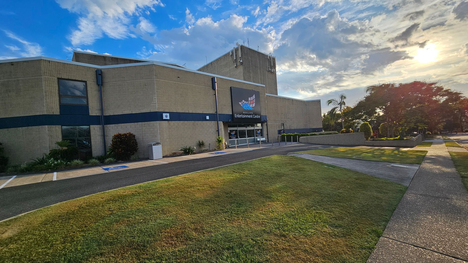 A large building with a beige exterior and a sign reading "Entertainment Centre" above the entrance. The entrance has glass doors and is flanked by small plants. To the right, a well-maintained lawn and a path lead to a garden area with trees and shrubs. The sun is low in the sky, casting long shadows on the ground. The sky is partly cloudy, and the overall setting appears calm and organised.