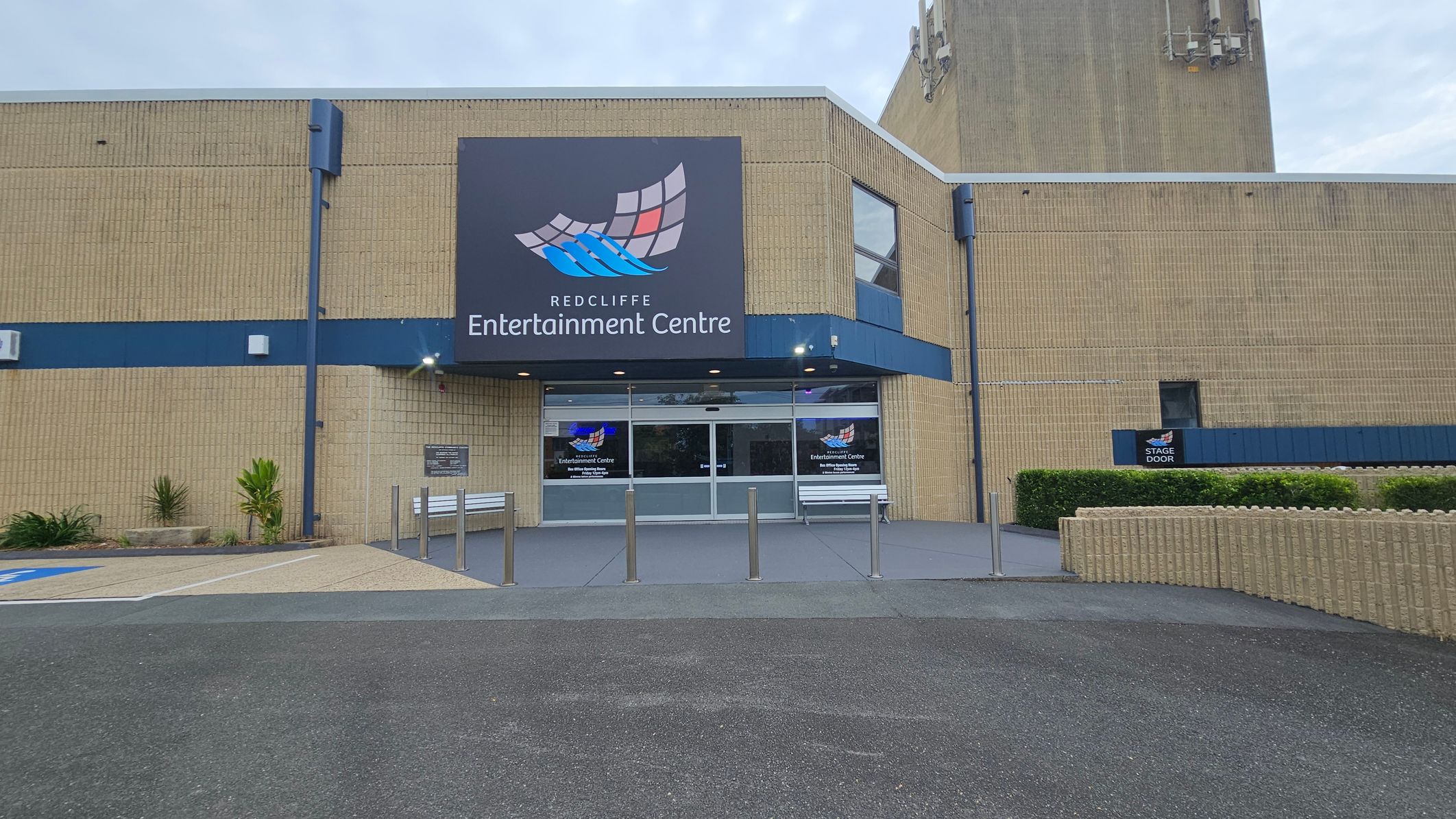 A brick building with a large sign reading "Redcliffe Entertainment Centre" above the entrance. The entrance has glass doors and is flanked by two white benches. There are several metal bollards in front of the entrance. To the left, there are small plants and a designated parking area. On the right, a sign indicates a "Stage Door" near a hedge. The ground is paved with asphalt and concrete, and the building has a blue trim.