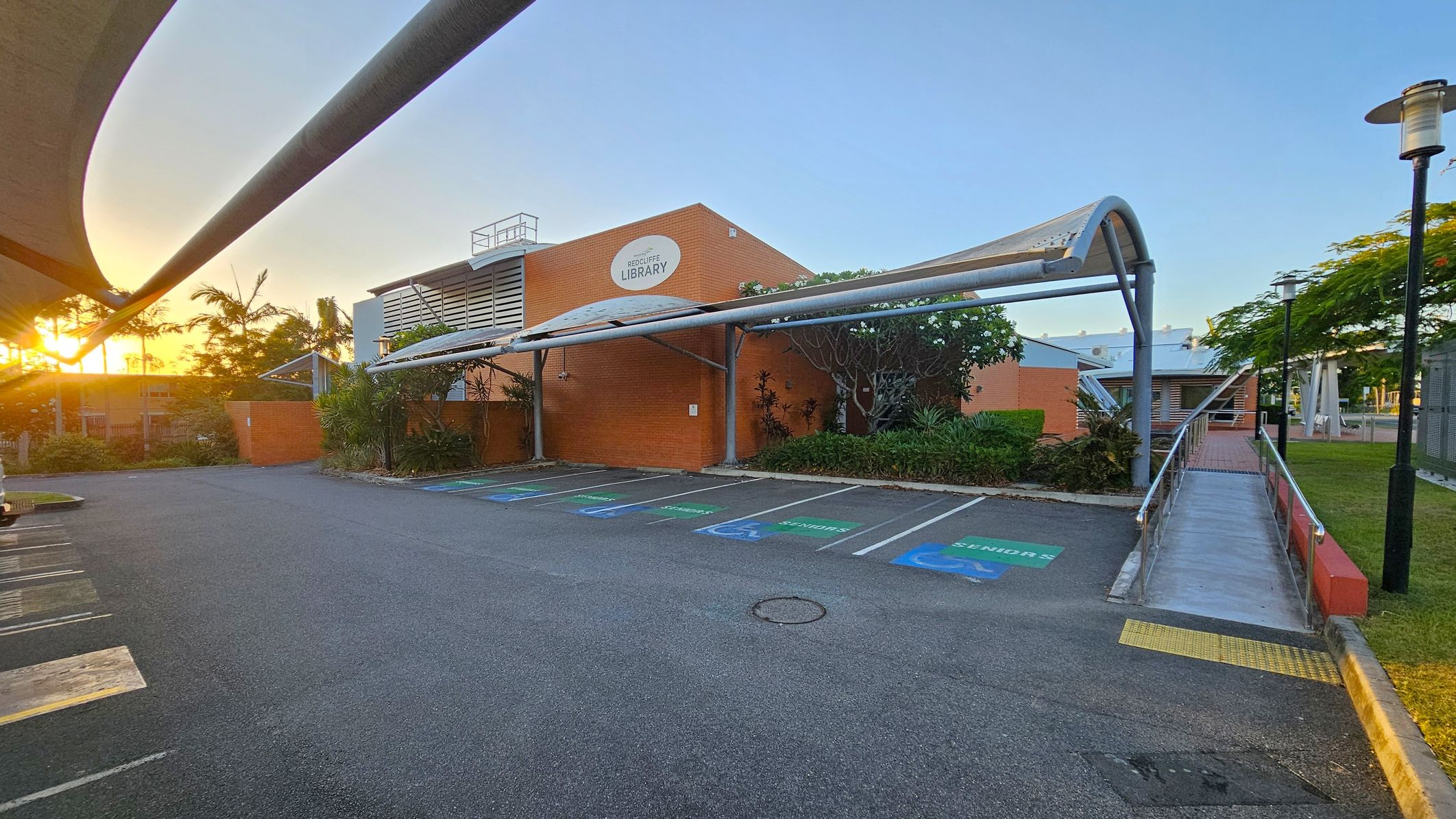 A parking area with designated spaces for seniors is in the foreground, marked with green and blue paint. A covered walkway leads to a brick building with a sign reading "Redcliffe Library" in the centre. The building is surrounded by greenery, including palm trees and shrubs. On the right, a ramp with metal railings leads to the entrance. The sun is setting on the left, casting a warm glow over the scene. A streetlamp is visible on the right, and the sky is clear.