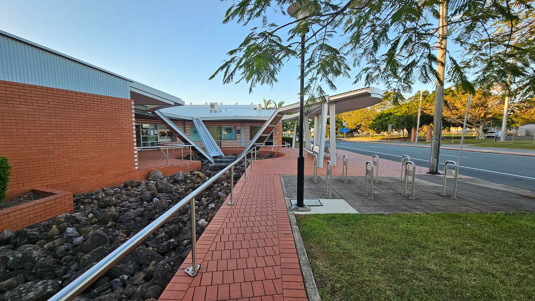 A brick pathway leads to a building with a red brick exterior on the left. The path is bordered by a metal railing and a bed of dark rocks. In the centre, a modern structure with angled metal beams and a glass entrance is visible. On the right, there are bike racks and a grassy area. A road runs parallel to the building, lined with trees and a utility pole. The sky is clear, and the area is well-lit.