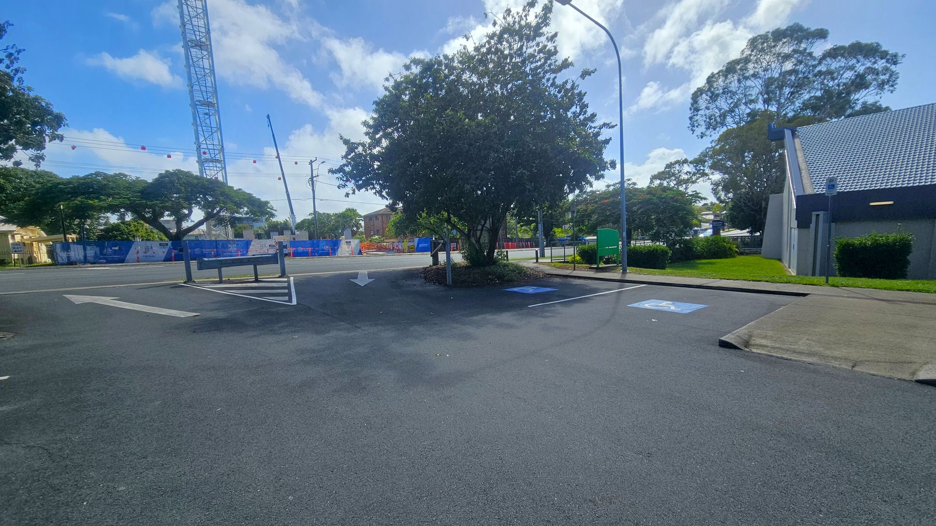 A car park with marked spaces, including a disabled parking spot on the right. A large tree is in the centre, surrounded by a small garden bed. To the right, a building with a grey tiled roof and a small grassy area. In the background, a construction site with a crane and blue fencing. Trees line the street, and a green bin is near the road. The sky is partly cloudy.