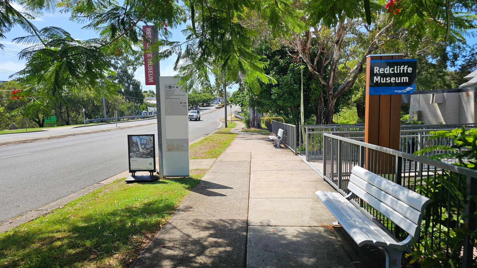 A footpath runs alongside a road, bordered by trees with green leaves and red flowers. On the left, a signpost and a small billboard are visible near the road. On the right, a sign for the Redcliffe Museum stands next to a metal railing and a white bench. The path is concrete, and the area is shaded by the trees above. The road curves slightly in the distance, with more trees and greenery visible further along.