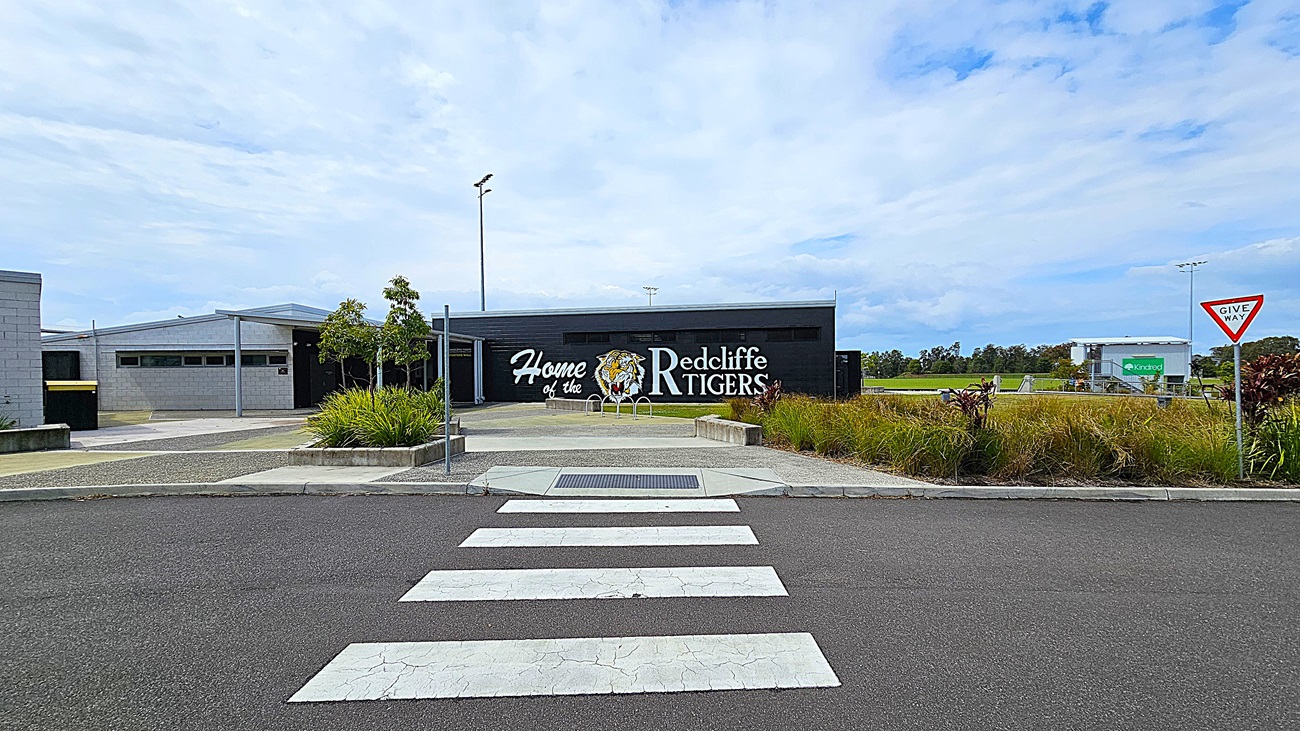 On the left, there is a grey building with large windows and a sign that reads "Home of the Redcliffe Tigers." To the right, a grassy area is visible, with some trees and shrubs. The foreground features a wide, asphalt road with a white pedestrian crossing marked by thick stripes. In the background, there are several light poles and a few signs, indicating parking areas.