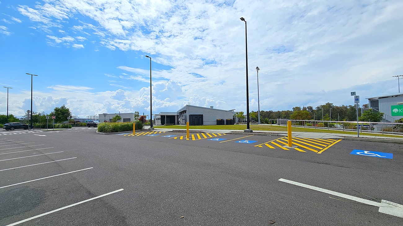 On the left, there are several empty parking spaces marked with white lines on a dark asphalt surface. A few light poles stand tall, providing illumination. To the right, a modern building with a flat roof features large windows and a black wall. In front of the building, there are designated parking spots for disabled access, marked with blue symbols and yellow stripes. The area is well-maintained, with some greenery visible in the background, including shrubs and trees.