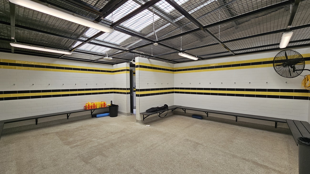 On the left, a black locker stands against a white tiled wall, with a round black bin positioned nearby. Above, a fan is mounted on the wall, and hooks are visible for hanging items. To the right, a row of beige lockers is set against the wall, with a whiteboard above a long bench. The floor is covered in a light-coloured carpet, providing a soft surface. The ceiling features a grid design with fluorescent lights, creating an open and well-lit environment suitable for changing and preparation.