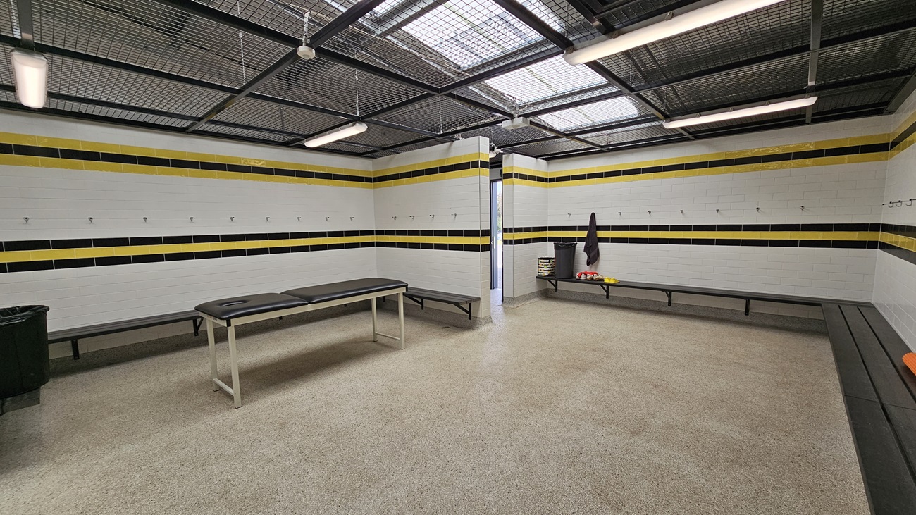 On the left, a long black bench runs along the wall, with several hooks above it for hanging items. Adjacent to it, a grey table is positioned in the centre of the room. On the right, a black bin stands next to a light grey door leading to another area. The walls are tiled with white tiles, featuring horizontal black and yellow stripes. The floor is a light speckled material, and the ceiling is a grid design with fluorescent lights and ventilation.