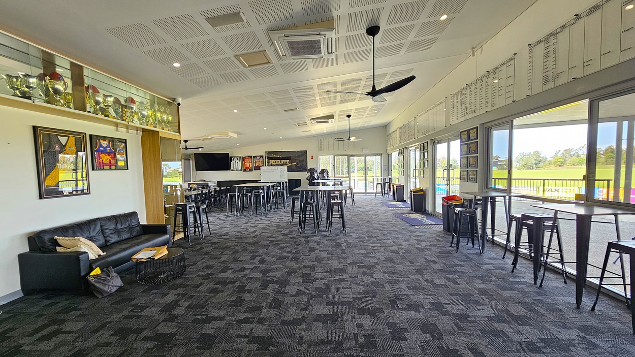 On the left, there is a glass door leading into a room with a dark carpet and tables visible inside. A welcome mat with a logo is placed just inside the entrance. To the right, a covered outdoor area features a smooth concrete floor supported by metal poles. In the background, there are green fields and a row of colourful advertising banners along a fence. The ceiling is flat and lined with light fixtures, providing illumination to the space.