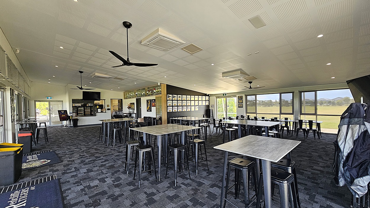 On the left, there are large glass doors leading outside, with a welcome mat in front. A bar area is visible, featuring shelves with drinks and a television mounted above. To the right, several tables with metal stools are arranged, and framed pictures hang on the wall. The floor is covered in dark patterned carpet, while the ceiling is flat with recessed lighting and ceiling fans.