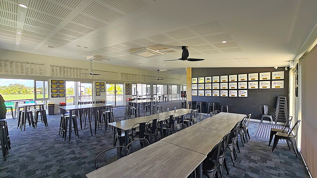 On the left, there are tall tables with metal stools arranged in a spacious area, with large glass doors leading outside. Above, the ceiling features recessed lighting and ceiling fans. To the right, a black wall displays framed certificates and images, while a stack of chairs is positioned nearby. The floor is covered in dark carpet with a textured pattern. The overall layout is open, allowing for easy movement between the tables and the entrance, creating a functional space for gatherings.