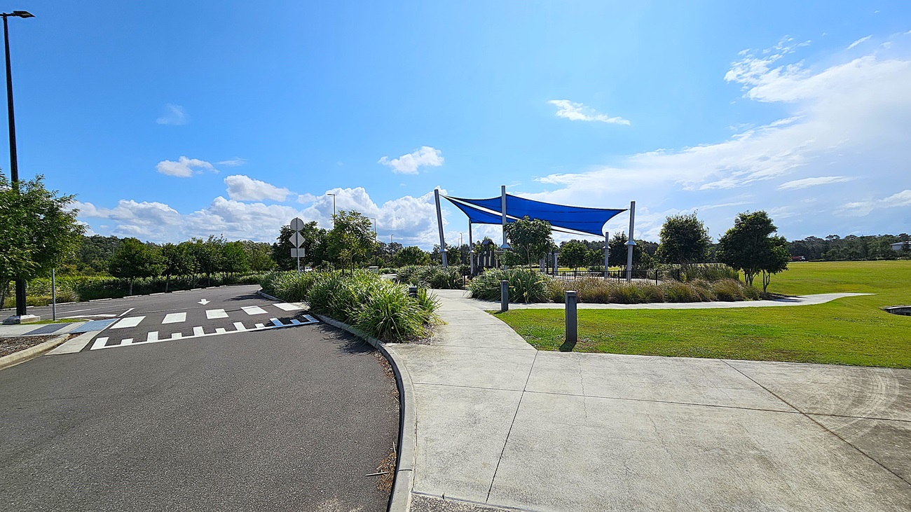 On the left, there are two large blue shade sails supported by tall silver poles, providing cover over a play area. Beneath the sails, a playground structure features a slide and climbing elements, surrounded by a soft, tan-coloured ground cover. To the right, a pathway leads towards a grassy field, with several trees in the background. The floor is made of smooth concrete, and the area is bordered by low fencing.