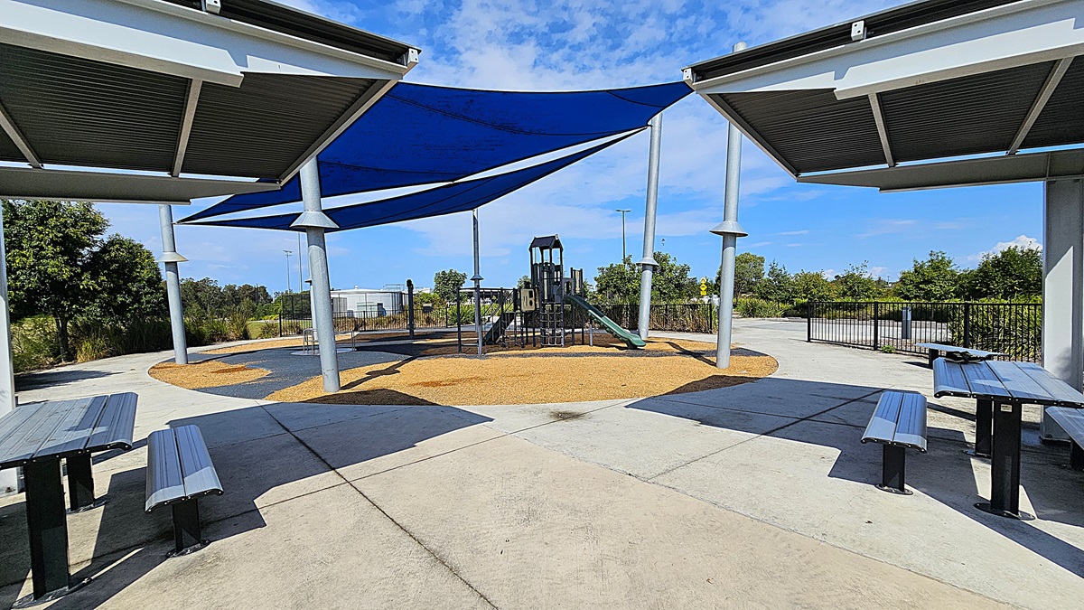 On the left, there are two metal picnic tables with attached benches, positioned on a smooth concrete surface. A blue shade sail is supported by silver poles above the tables. In the centre, a playground structure features a slide and climbing elements, surrounded by a soft, tan-coloured ground cover. To the right, the pathway curves away, leading to a fenced area with greenery. The floor is primarily concrete, and the ceiling consists of the shade sail overhead.