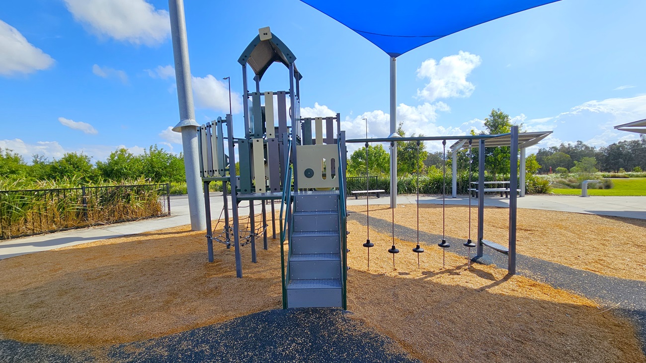 On the left, there is a multi-level playground structure with a slide and climbing features, accessed by a set of grey stairs. The ground around the structure is covered in tan wood chips, providing a soft landing. To the right, a swing set with several swings hangs from a metal frame, also situated on the wood chip surface. The area is shaded by a large blue shade sail supported by tall poles. The surrounding space includes green shrubs and trees, with a paved pathway visible in the background.