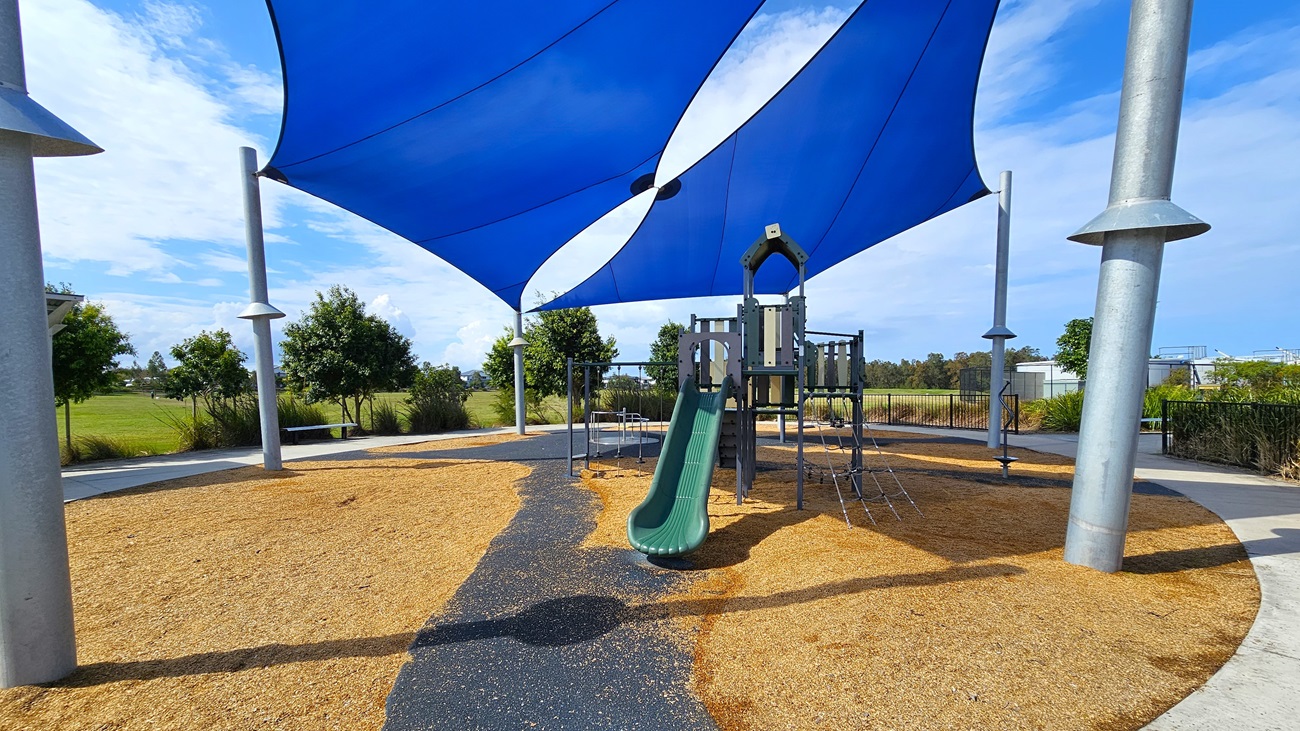 On the left, a green slide is attached to a multi-level playground structure, which includes climbing features and a small platform. The ground is covered with tan wood chips, providing a soft surface for play. To the right, there are several metal poles supporting a large blue shade sail that covers the play area. In the background, there are low shrubs and trees, with a paved pathway visible. The floor is primarily wood chips, and the ceiling consists of the shade sail.
