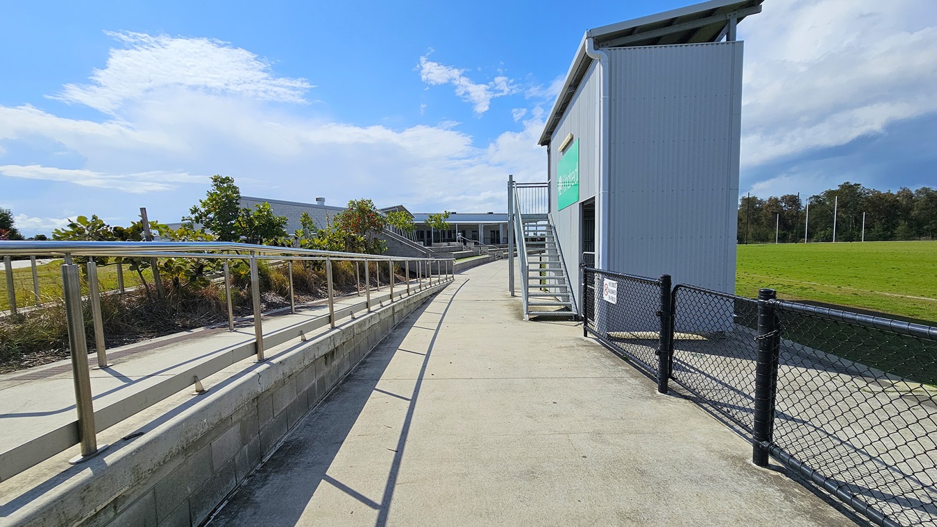 On the left, a gently sloping concrete ramp with a metal handrail leads towards a grassy area, bordered by low shrubs. The surface is smooth and grey, extending towards a building in the background. To the right, a set of metal stairs ascends to a higher level of the building, which has a grey exterior and a green sign. A black chain-link fence runs along the pathway, separating it from the adjacent field.