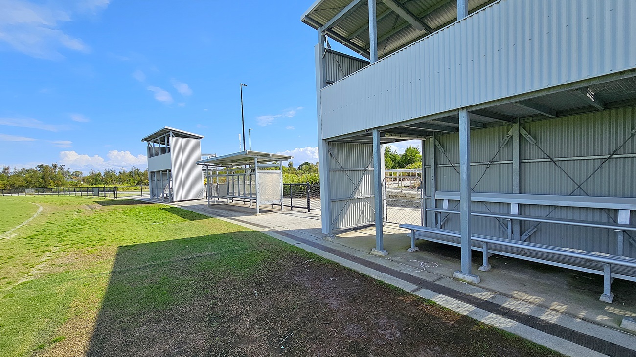 On the left, a tall, grey structure with large windows serves as a viewing tower, providing an elevated perspective of the field. Adjacent to it, a covered seating area with metal benches is visible, positioned along a concrete pathway. To the right, another grey structure features a similar design, with open sides and a roof, offering additional seating. The ground is a mix of grass and bare earth, with a well-maintained grassy area extending into the background, bordered by a black fence.
