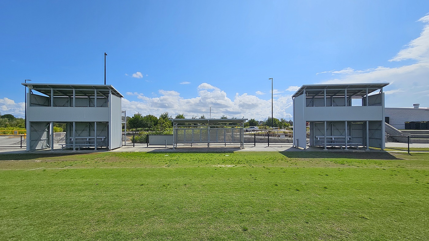 On the left, a grey metal structure features a covered viewing area with benches, supported by vertical posts. Adjacent to it, a similar structure is visible on the right, both facing a grassy field. In the middle, a concrete pathway runs between the two structures, leading towards a fenced area in the background. The floor is primarily grass, with some bare patches, and the ceiling is flat and made of metal, providing shelter for spectators.