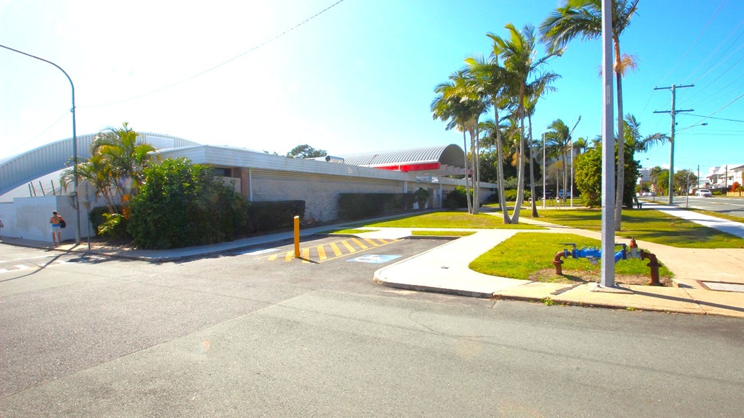 At the left side of the Redcliffe War Memorial Swimming Pool building, there is an accessible car parking space, demonstrated by the road marking painted with yellow lines and 2 wheelchair icons. Water valves are visible at the right corner alongside the walkways. There are also hedges along the sidewalk that are leaning against the long brick wall of the building at the left corner of the accessible car space.