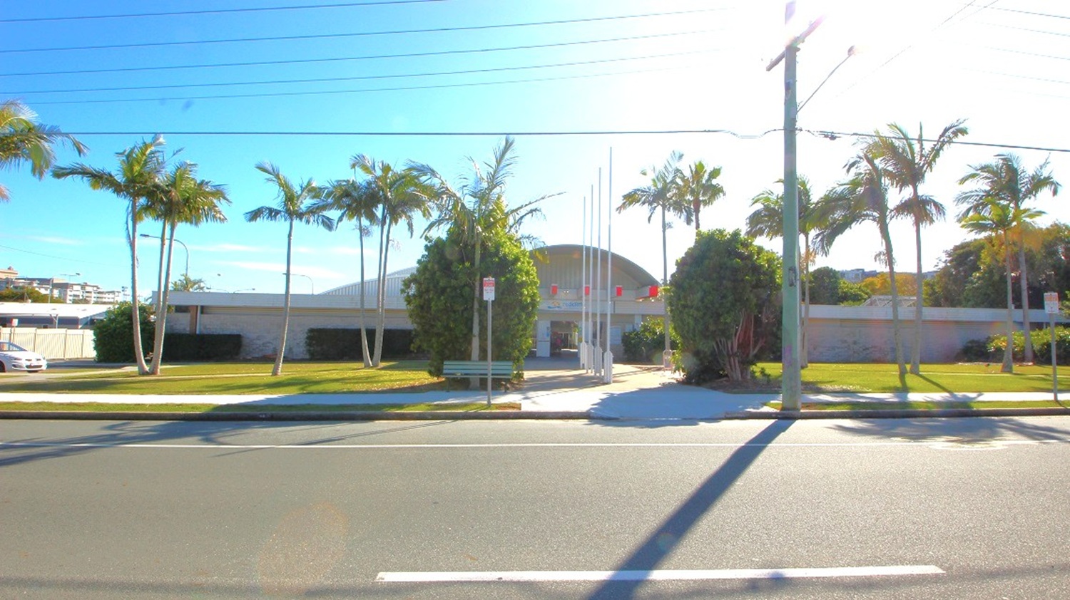The Redcliffe War Memorial Swimming Pool is close to Sydney Street and is landscaped with rustling palm trees, one of which has a bench underneath it, shade-giving hedge plants, lush green grasses, and four white flagpoles lined up. The building entrance is symmetrically placed in the middle of the two separate front long walls made of bricks.