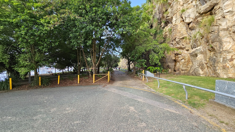 To the left is an accessible car park with a grey vehicle facing the river. There are three red silhouettes of a human with outstretched arms holding various structures overhead, with rubbish bins positioned nearby. Kangaroo Point Cliffs are on the right, bordered by metal railings. Skyscrapers are visible further ahead, with trees and grass surrounding the area.