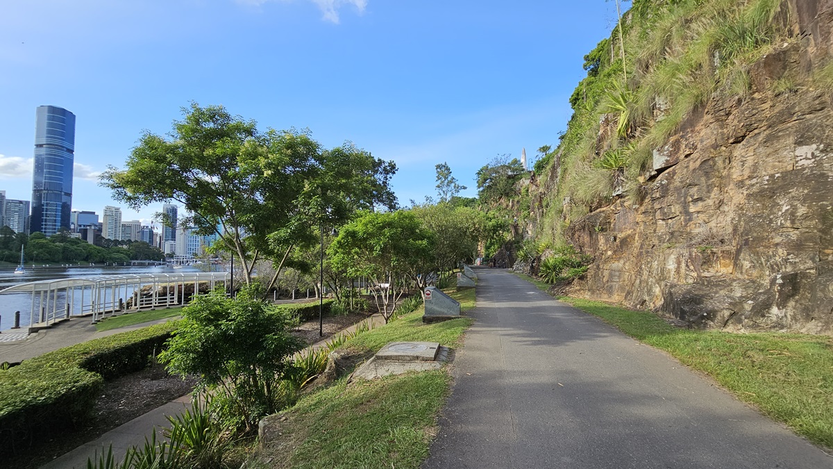 A straight road is bordered by a cliff on the right and trees and solid structures with lighting on the left. To the left of the road is lower ground with landscaped plants and neatly trimmed shrubs. Shade structures are positioned near the lake. Further ahead, tall skyscrapers are visible.