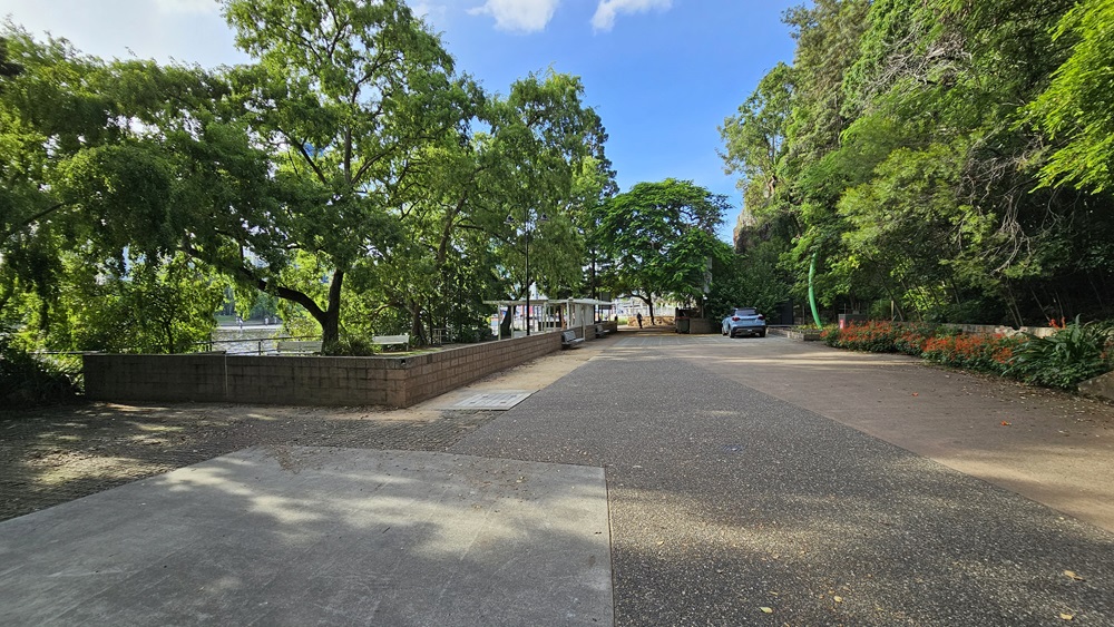 The road is lined with trees and flowering plants and shrubs on the right. On the left, there is a river and a park with a shade structure, and bench chairs, enclosed by a low concrete fence. A grey vehicle is parked on the right side of the road.