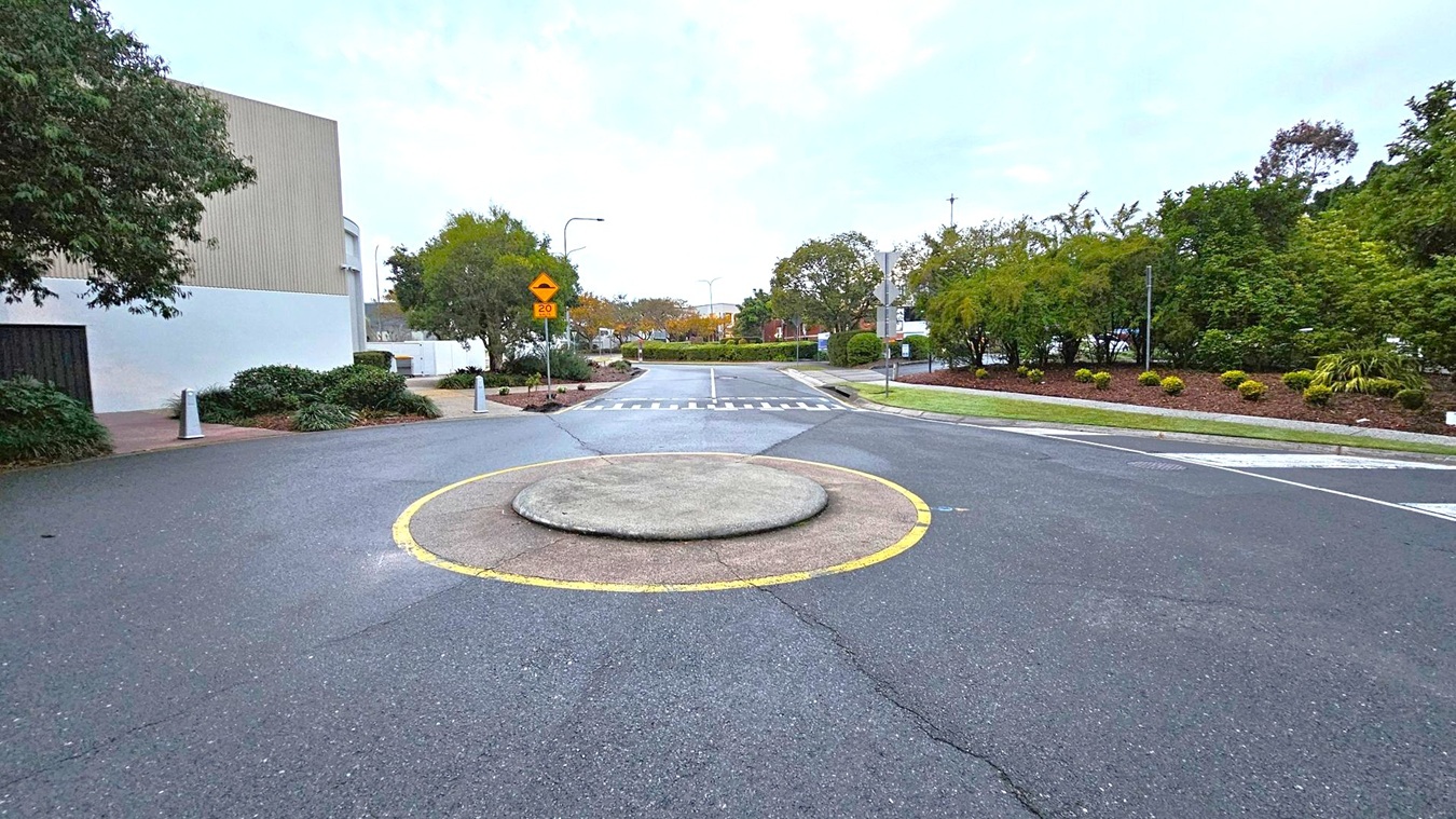A small roundabout is in the centre of a road, marked by a circular concrete island with a yellow border. On the left, there is a large building with a beige and white facade, accompanied by some greenery. On the right, there are well-maintained bushes and trees, with a grassy area. In the background, more trees and a few street signs are visible, along with a pedestrian crossing. The road surface is dark grey with visible markings.