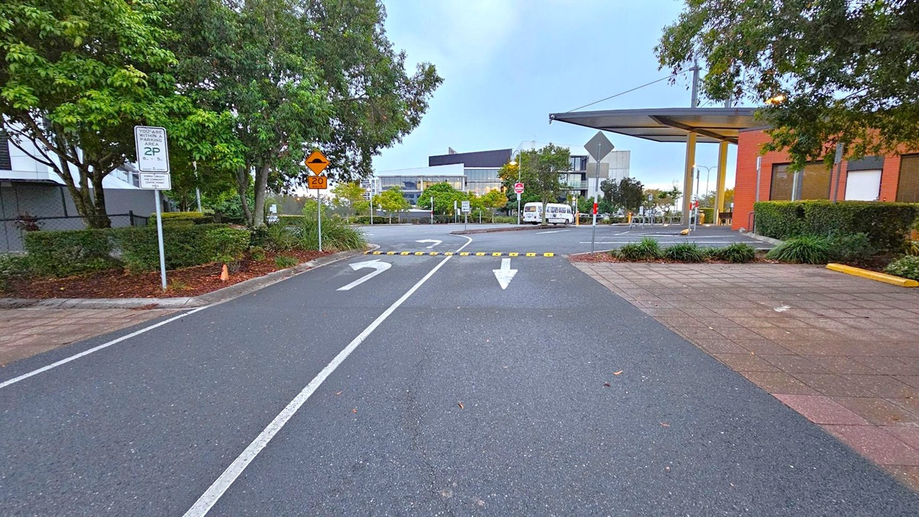 A road with two white arrows painted on the surface, one pointing straight and the other curving left. A speed bump with yellow markings is visible. On the left, there are trees and a parking sign. On the right, a building with an overhang and more trees. In the background, a few buildings and a parked van are visible. The pavement is bordered by a tiled walkway on the right.