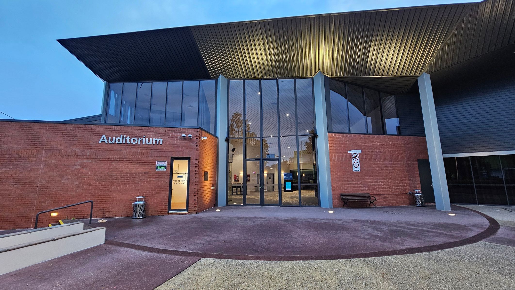 A modern building with a large glass entrance in the centre, flanked by red brick walls. The word "Auditorium" is displayed on the left wall. A small door with a sign is on the left, and a bench is on the right. The roof is angular and metallic, extending over the entrance. The ground is a mix of concrete and textured surfaces, with a ramp and steps on the left. The interior is visible through the glass, showing a lit hallway.
