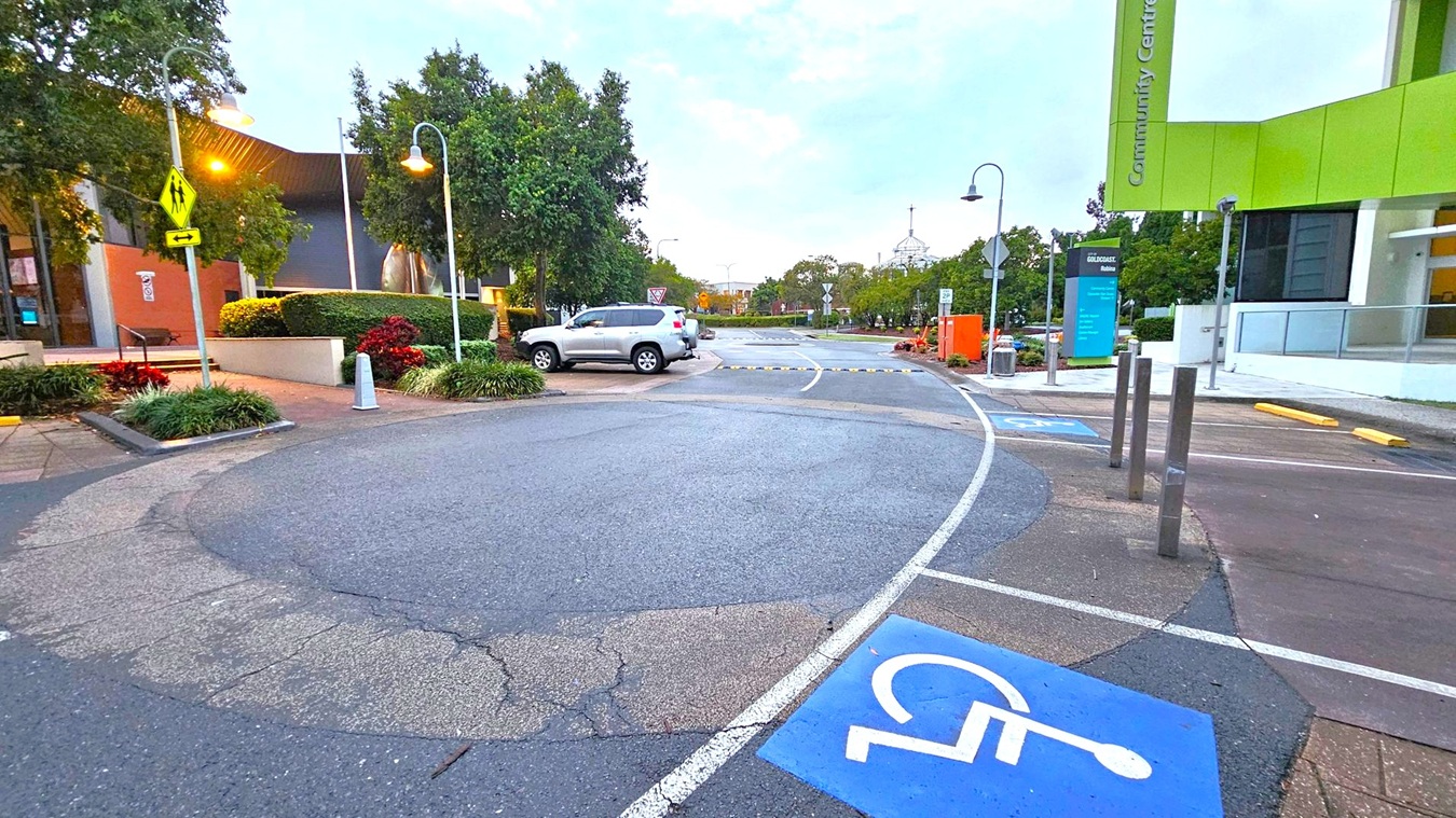 A street scene with a focus on a parking area. In the foreground, a blue disabled parking space is marked on the asphalt. On the left, there are trees and a building with a brown and grey facade. A silver SUV is parked nearby. On the right, a modern building with a green sign reading "Community Centre" is visible. Street lamps line the road, and there are various signs and bollards. The background features more trees and a clear sky.