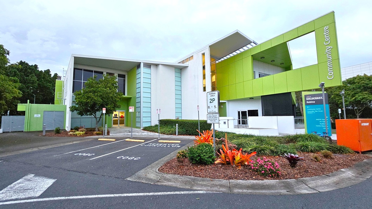 A modern building with a green and white facade, labelled "Community Centre" on the right. The structure features large windows and a prominent overhang. In the foreground, there is a small garden with vibrant plants and a parking area with marked spaces. An orange utility box is visible on the right, and a sign provides parking information. Trees and shrubs are present on the left, adding greenery to the scene.