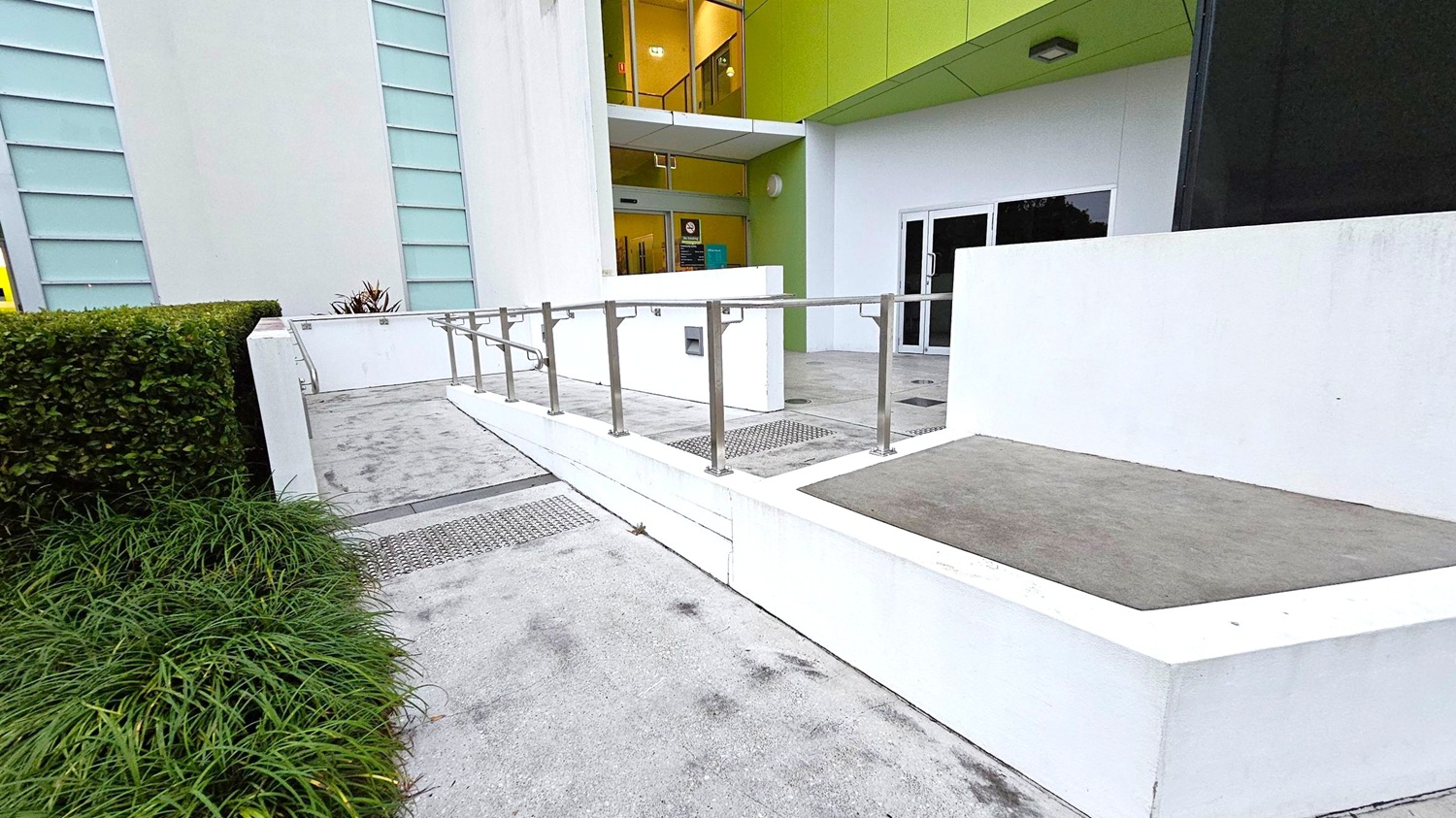 A concrete ramp with metal railings leads to a building entrance. On the left, there are green bushes and grass along the path. The building has a white facade with vertical blue-tinted windows. The entrance features glass doors and a green overhang above. Inside, a yellow-lit interior is visible through the glass. The ground is paved with concrete, and there are metal grates near the ramp.