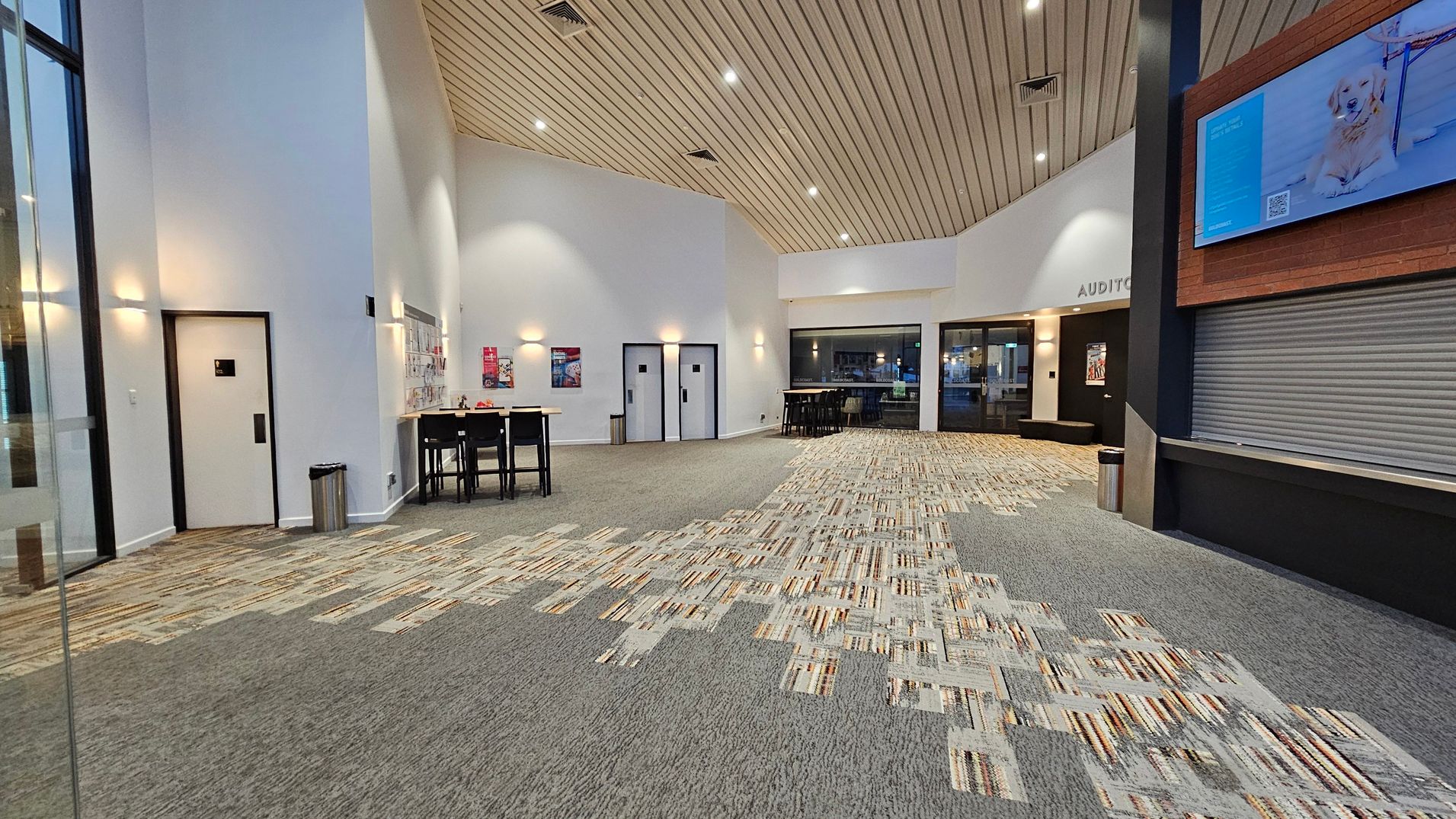 A spacious lobby with a patterned carpet floor in shades of grey and beige. On the left, there are white walls with a door and a small table with chairs. The ceiling is wooden with recessed lighting. In the centre, two doors are visible, and a high table with stools is placed nearby. On the right, a large screen is mounted above a closed shutter, and a sign reads "AUDITORIUM" above glass doors. A waste bin is positioned near the shutter.