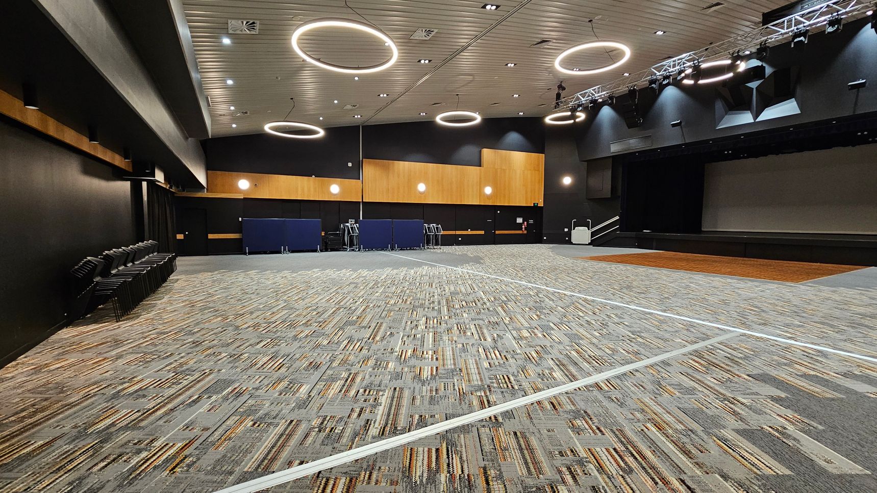A large, modern auditorium with a patterned carpeted floor. On the left, there are rows of stacked chairs against a dark wall. The ceiling features circular light fixtures and a grid of spotlights. The back wall has wooden panels and small round lights. On the right, a stage with a wooden floor section and steps leading up to it. The overall colour scheme is neutral with dark and wooden accents.