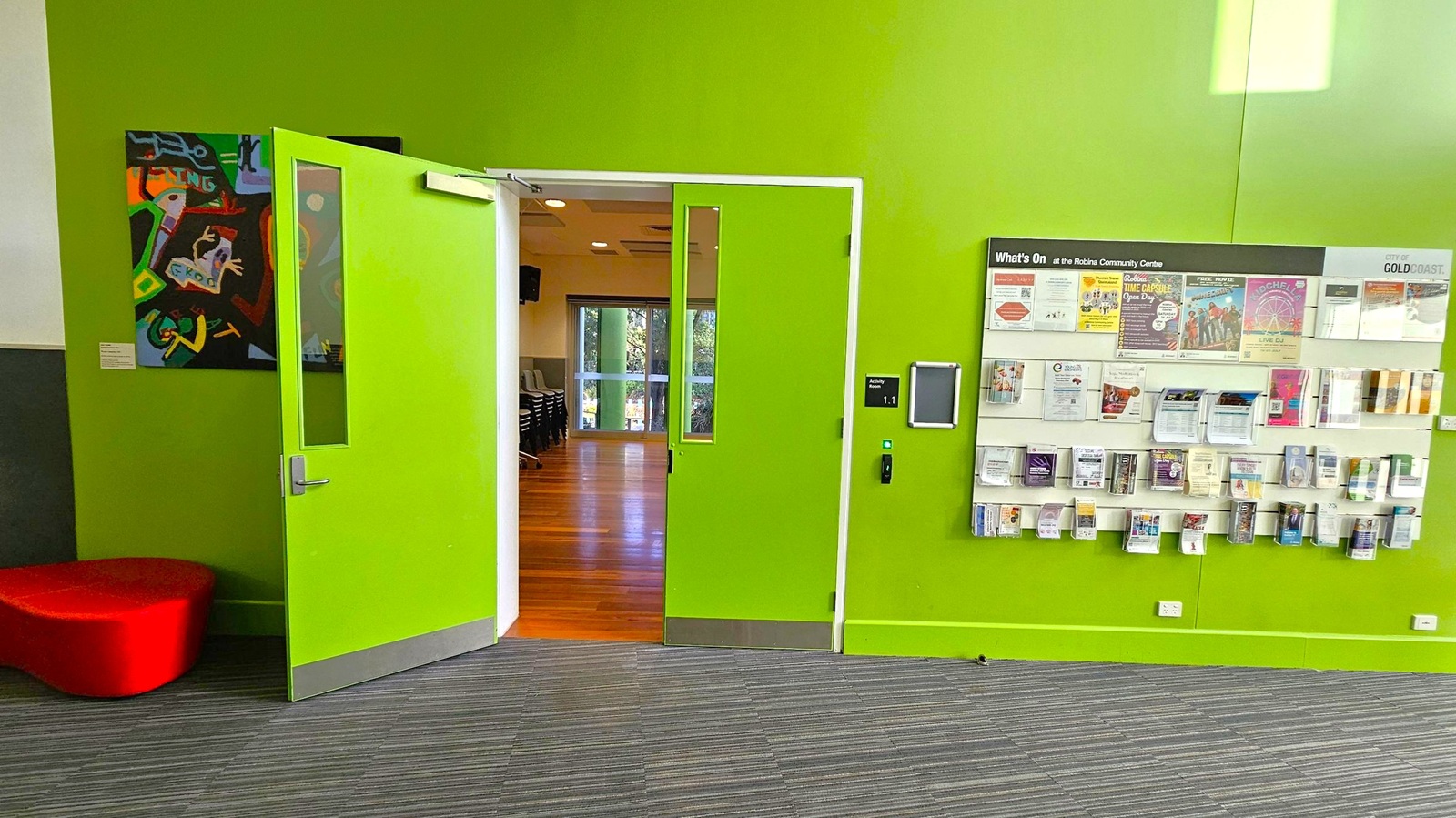 A room with bright green walls and a partially open door leading to another room with wooden flooring. On the left, there's a colourful abstract painting and a red, rounded seat. On the right, a wall-mounted display holds various brochures and flyers under a "What's On" sign. The floor is covered with grey striped carpet.