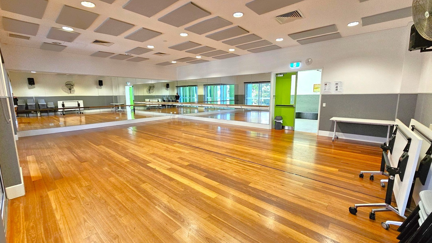A spacious room with a polished wooden floor and a ceiling featuring recessed lighting and acoustic panels. The left wall is lined with large mirrors, reflecting the room's interior. On the right, there are stacked chairs and foldable tables. The back wall has a green door and large windows with vertical blinds. A small table and a bin are near the door. The room appears to be a dance or exercise studio.