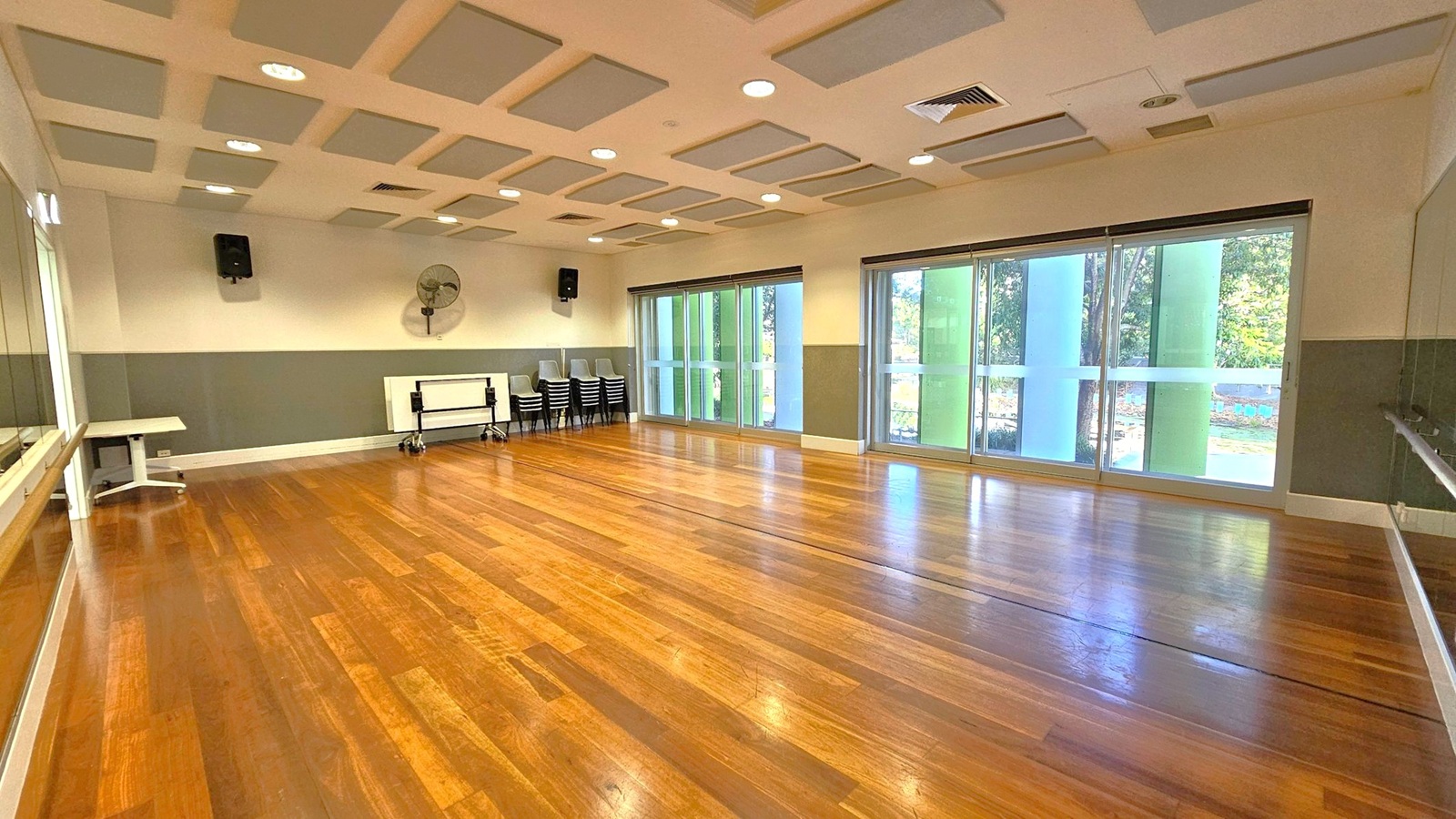 A spacious room with polished wooden floors and a ceiling featuring square acoustic panels. On the right, large windows with vertical green and blue panels allow natural light to enter. The left side has a long mirror and a white table. At the back, stacked chairs and a folded table are against the wall, with a wall-mounted fan and speaker above. The walls are painted in a two-tone scheme, with the lower half in grey.