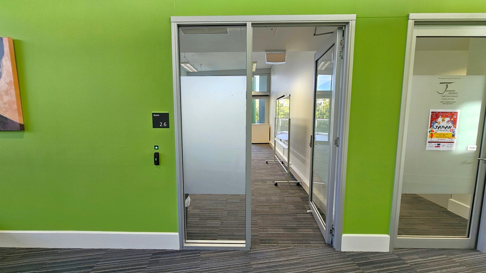 A room with a green wall and a glass door labelled "2.6" on the left. The door has a frosted lower section and a clear upper section. Inside, there's a carpeted floor with a striped pattern and a window at the far end. To the right, another glass door with a poster on it. A portion of a painting is visible on the far left.
