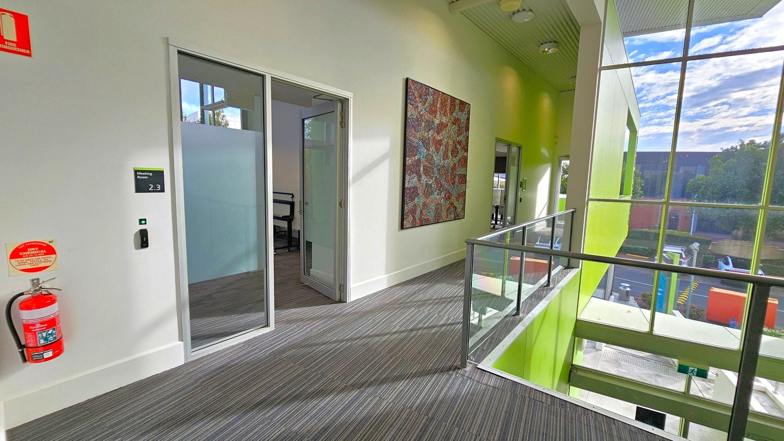 A corridor with grey carpeted flooring and a white ceiling. On the left, there's a glass door labelled "Meeting Room 2.3" with a fire extinguisher mounted on the wall. A colourful abstract painting hangs on the wall further down. On the right, large windows with green accents provide a view of the outside, showing trees and a building. A glass railing lines the edge of the corridor, overlooking a lower level.
