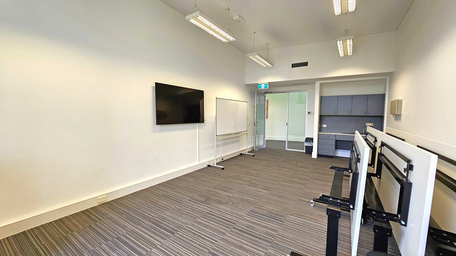 A room with a striped carpeted floor and white walls. On the left, a flat-screen TV is mounted on the wall next to a whiteboard on wheels. The ceiling has several rectangular fluorescent lights. On the right, there are stacked tables with black metal legs. In the background, a doorway with a glass door leads to another room, and there are grey cabinets and a small bin near the wall.