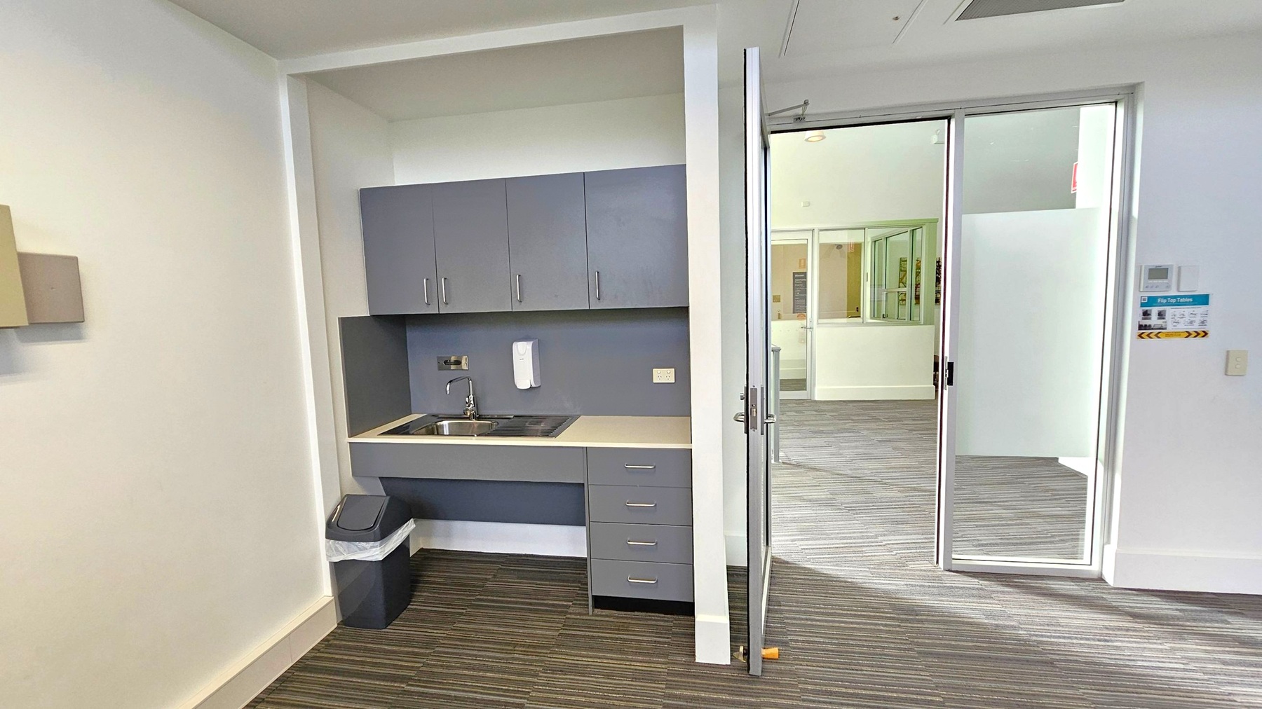 A small kitchenette is on the left with grey cabinets, a sink, and a countertop. Below the sink is a bin. The floor has a striped carpet. On the right, a glass sliding door leads to another room with a similar carpet. The walls are white, and there is a sign with instructions near the door. The ceiling is plain with a vent visible.