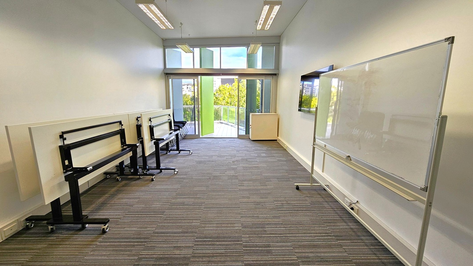 A room with a striped carpeted floor and a high ceiling with fluorescent lights. On the left, there are several foldable tables stacked against the wall. On the right, a large whiteboard on wheels is positioned near the wall. At the far end, large windows and a glass door open to a balcony, revealing green trees outside. A small cabinet is placed near the windows.